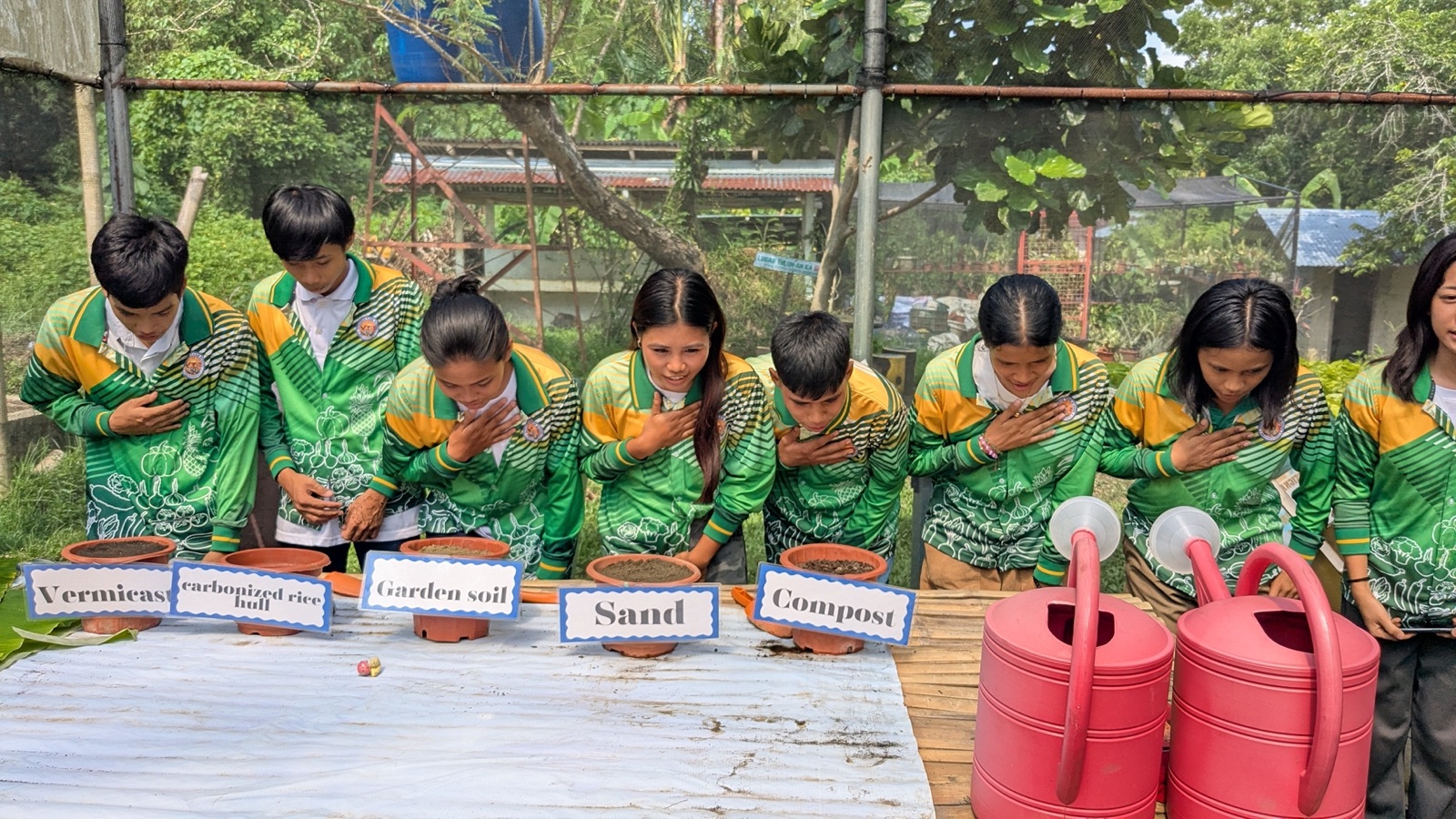 Students in green uniforms with hands over their hearts at a soil-science station.