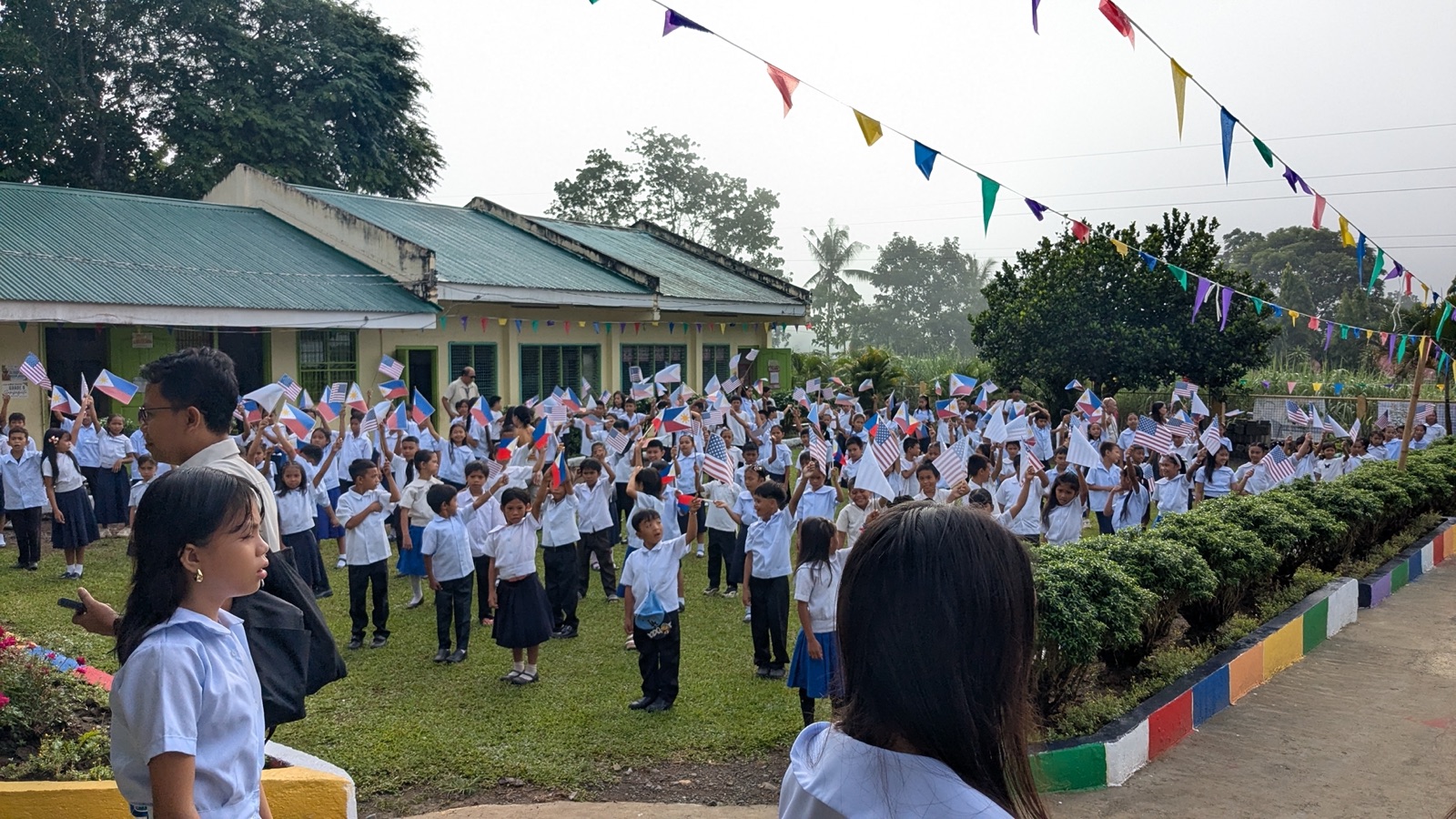 Students waving Philippine and U.S. flags at an outdoor school assembly.