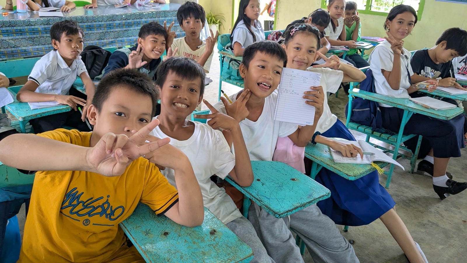 Students at their desks smiling and giving peace signs in class.