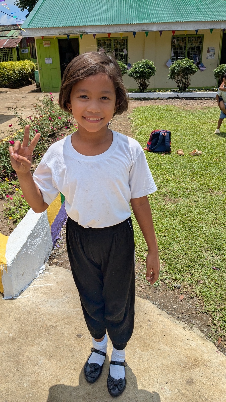 A young student giving a peace sign on a path outside her school.