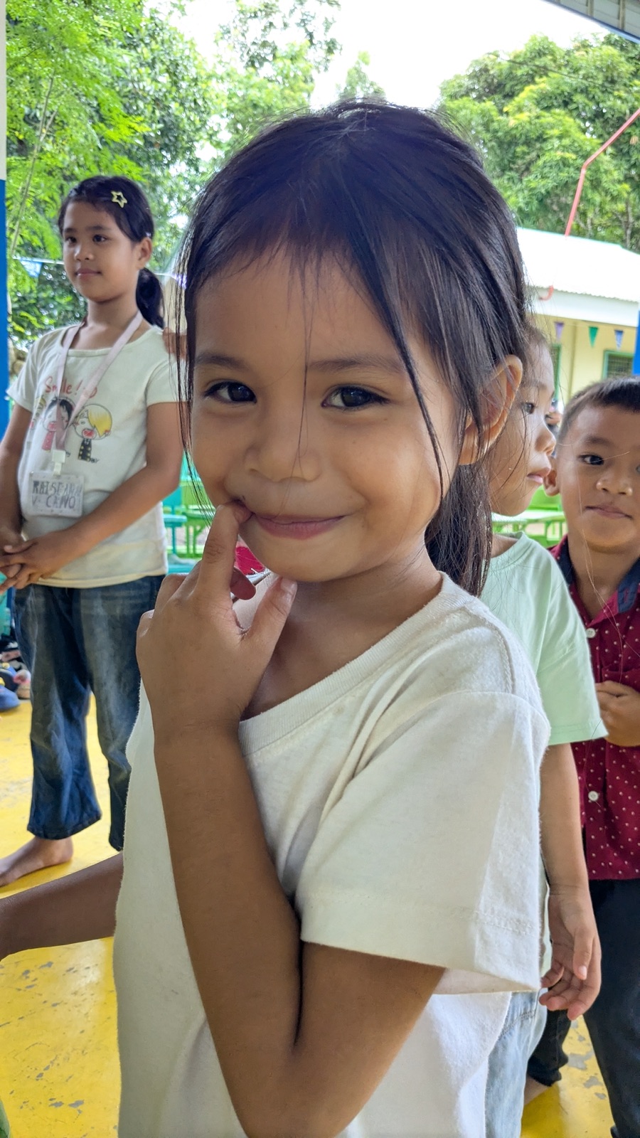 A young girl smiling shyly among classmates at a covered court.