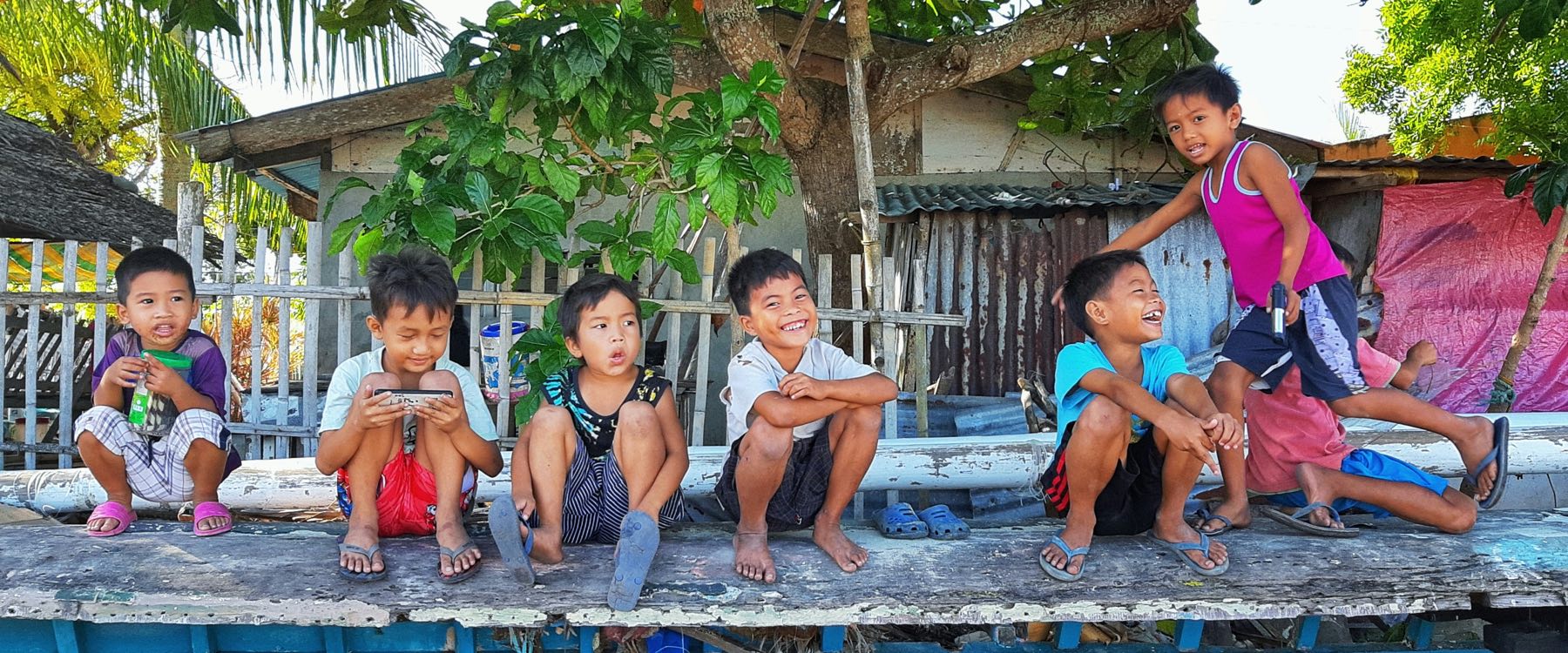 Seven children sitting in a row along a plank beside a boat, under a tree.