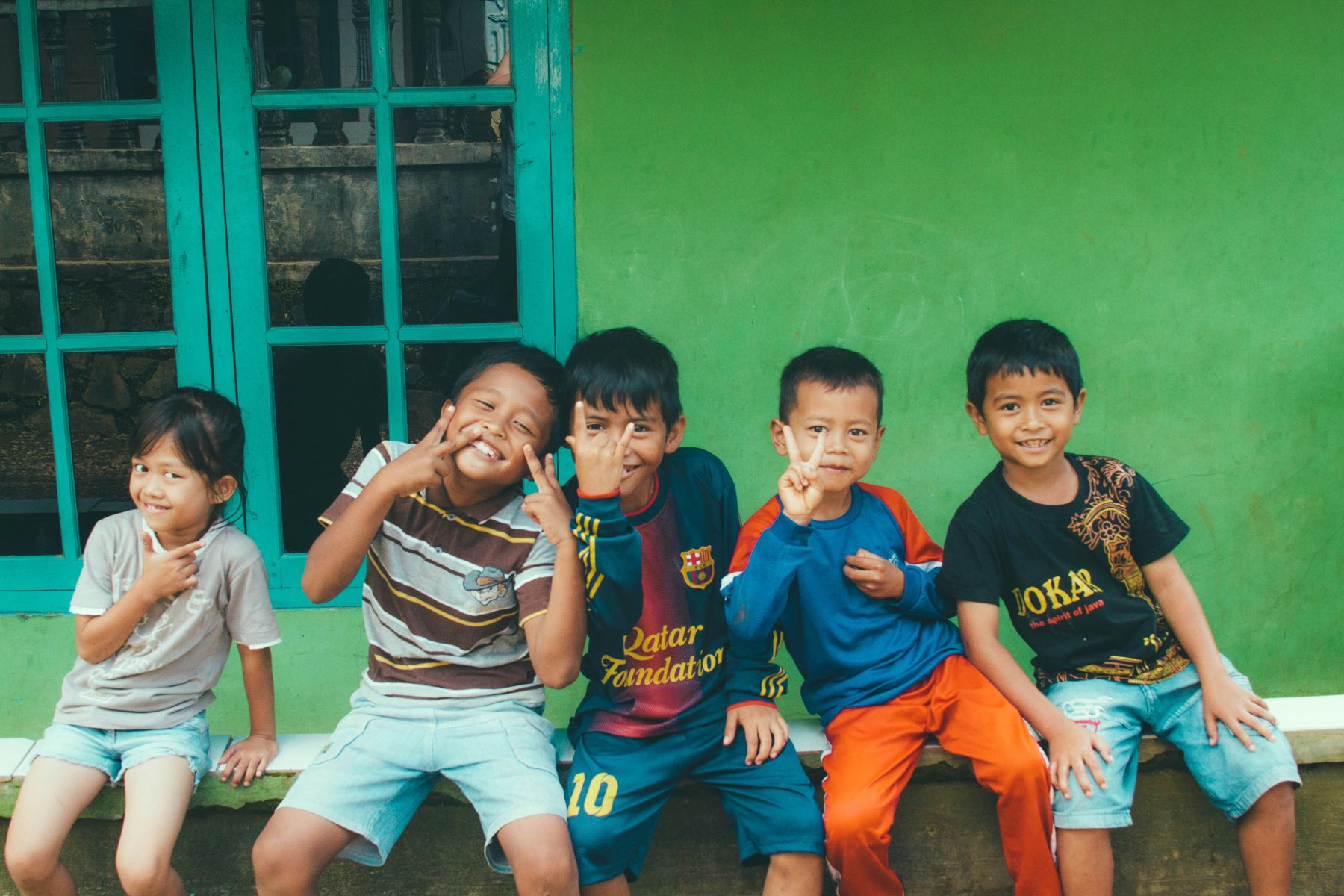 Five children sitting in a row against a bright green wall, smiling and making peace signs at the camera.